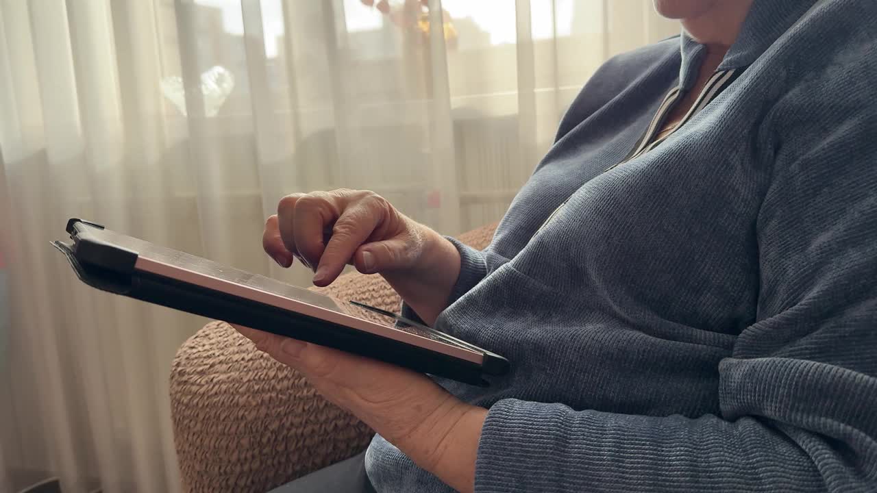 一位时髦的老妇人坐在扶手椅上，在家里用智能平板电脑查找重要信息视频下载