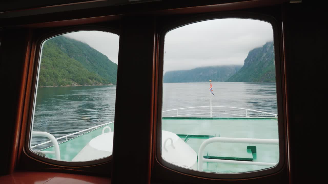 透过船的舷窗，可以看到风景如画的挪威峡湾。在前面你可以看到船的机头和挪威国旗视频下载