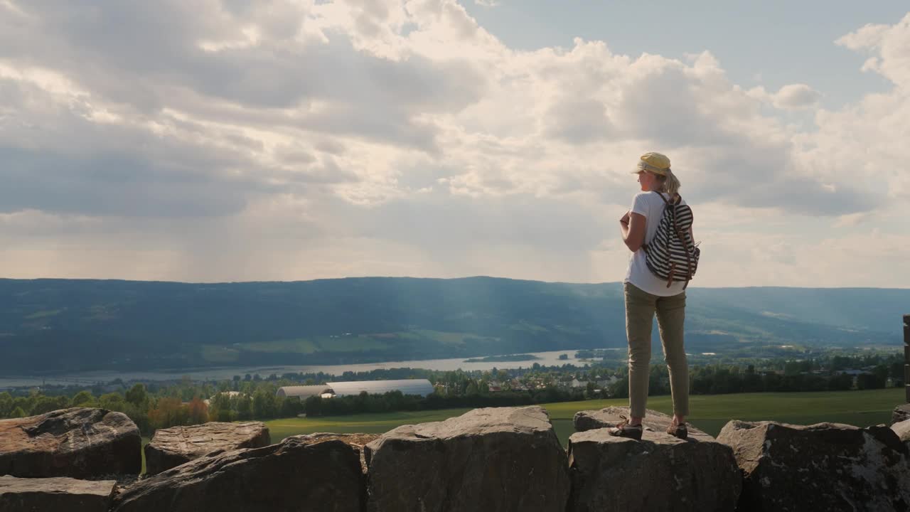 一个背着背包的女人站在一堵古老的石墙上，望着远处引人注目的天空和地平线视频下载