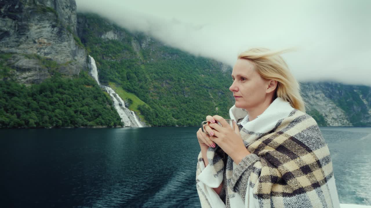 船上的一位女乘客一边喝着热茶，一边欣赏着挪威峡湾的美景视频下载