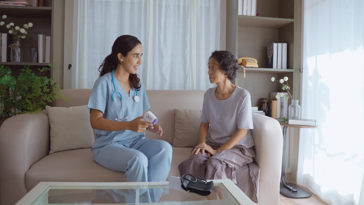 拉丁美洲的一名女护士和一名泰国老年女病人一起坐在沙发上，穿着休闲服，用数字体温计测量病人的体温，然后向病人解释健康检查的结果。视频下载