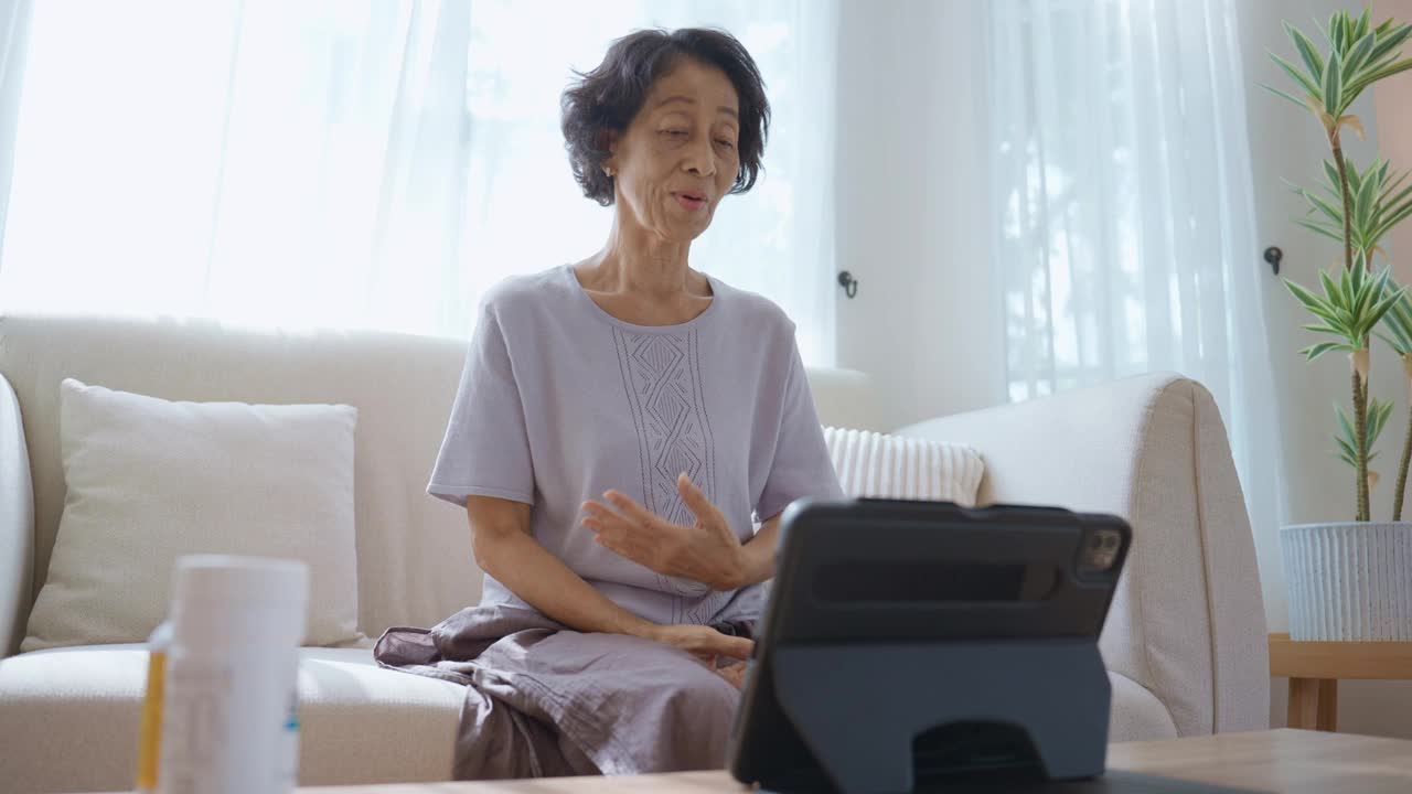 在一张特写照片中，一位泰国老年妇女穿着休闲服，坐在舒适客厅的沙发上，通过她的电子平板电脑与医生交谈，以更新她的健康状况，以预防疾病。视频下载