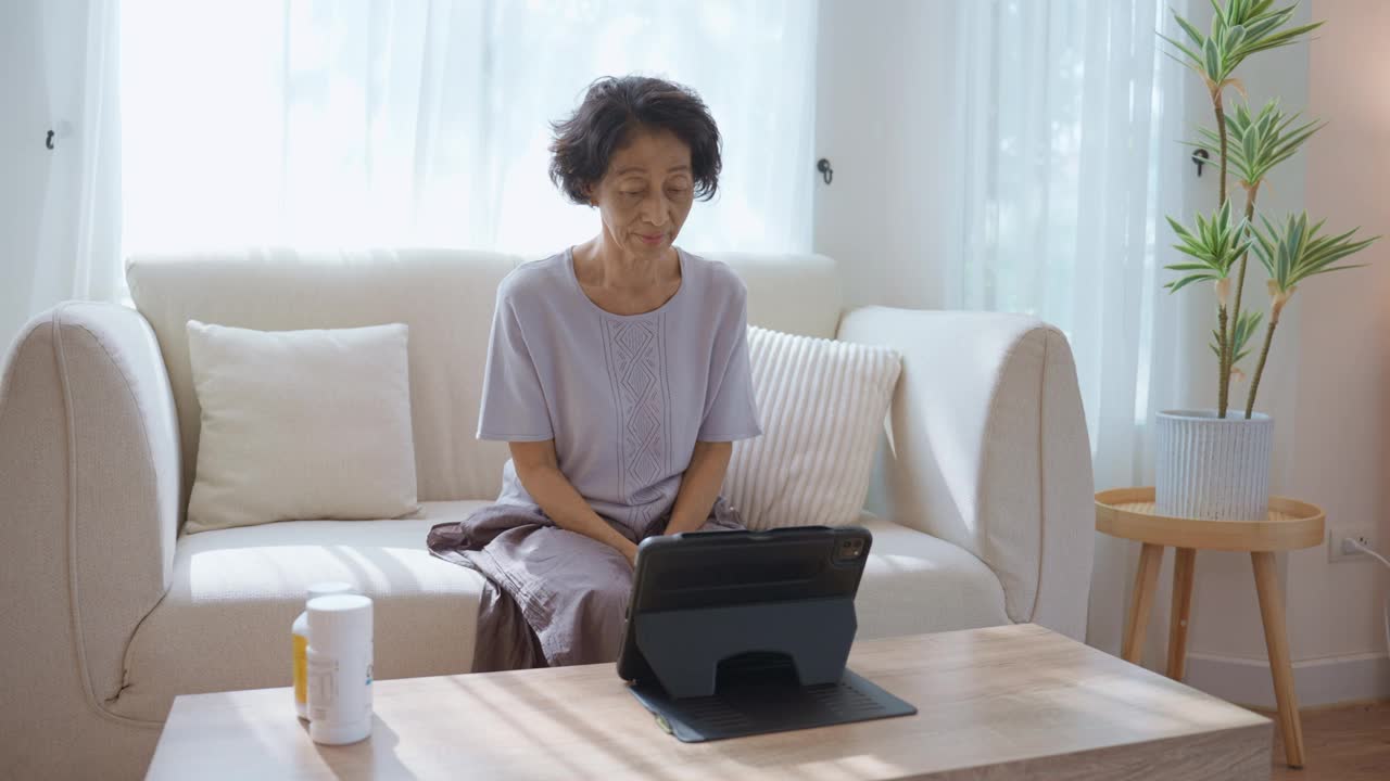 一位泰国老年女性坐在舒适家中的沙发上，通过她的平板电脑向在线医生描述她的症状和疾病，以更新她的身体健康状况。视频下载