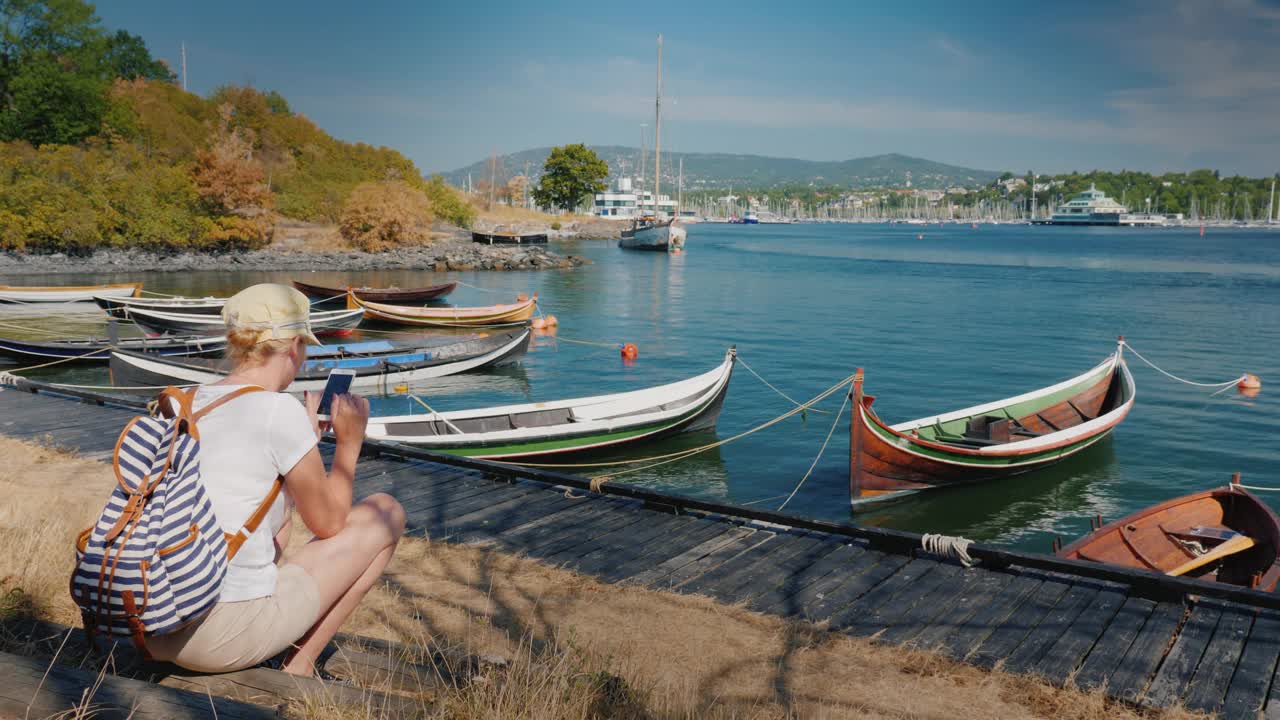 在挪威奥斯陆市的海滨，一名背着背包的妇女坐在渔船附近。使用智能手机。去斯堪的纳维亚国家旅行视频下载