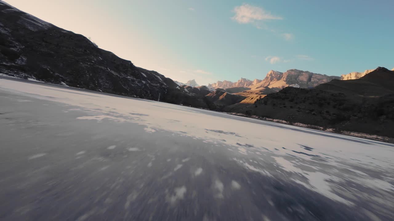 冬季，从高处俯瞰湖面结冰的水面，一架运动FPV无人机飞过湖面。背景是一座白雪皑皑的山。这个季节游客们来这里滑冰视频下载