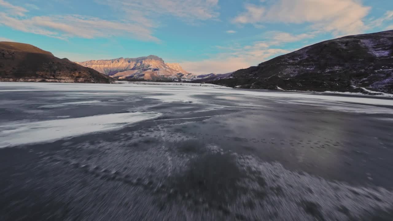 快速的FPV无人机在冰冻的湖面上飞行，背景是白雪皑皑的日落山脉视频下载