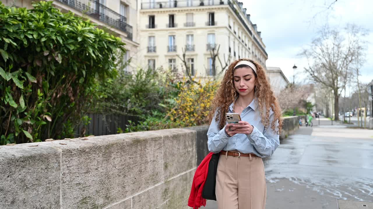 微笑的Z世代卷曲女性在巴黎使用智能手机视频下载