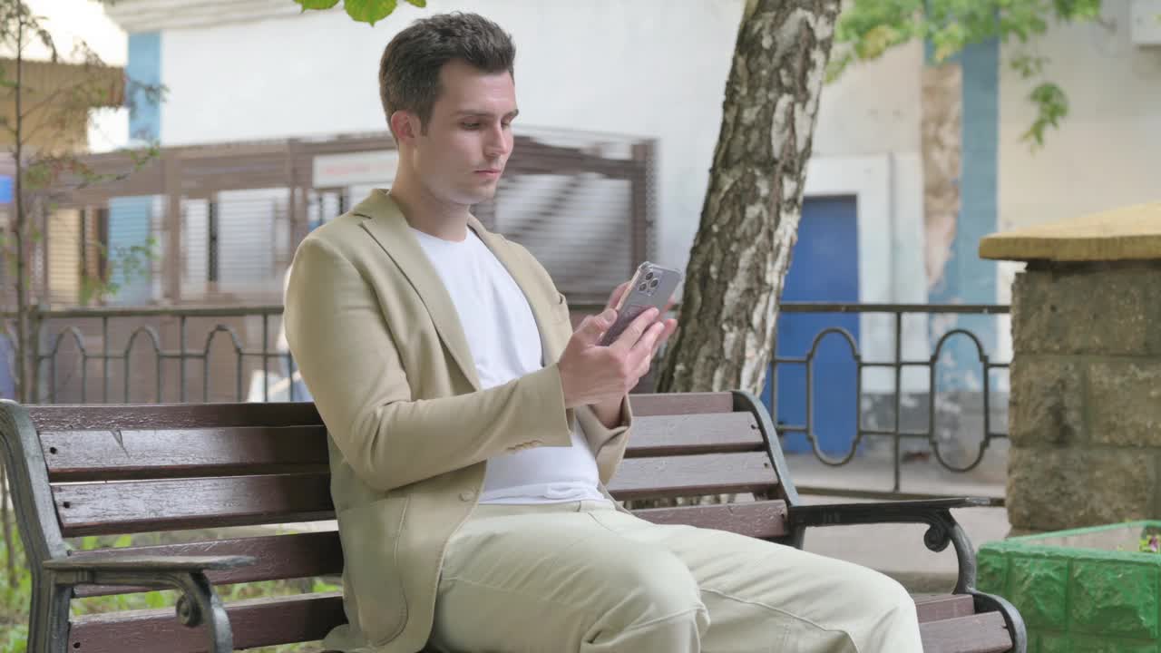 一个年轻人坐在户外长凳上玩智能手机庆祝成功视频下载