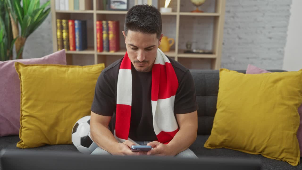 一个精力充沛、留着胡子、戴着围巾的西班牙裔年轻人坐在家里，手里拿着智能手机，前面放着一个足球和一台笔记本电脑。视频下载