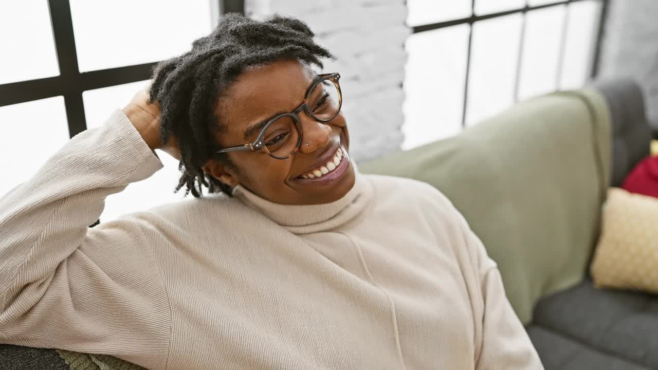 一位戴着眼镜、梳着脏辫、心情愉快的非裔美国妇女在室内的沙发上放松，对着砖墙的背景微笑着。视频下载