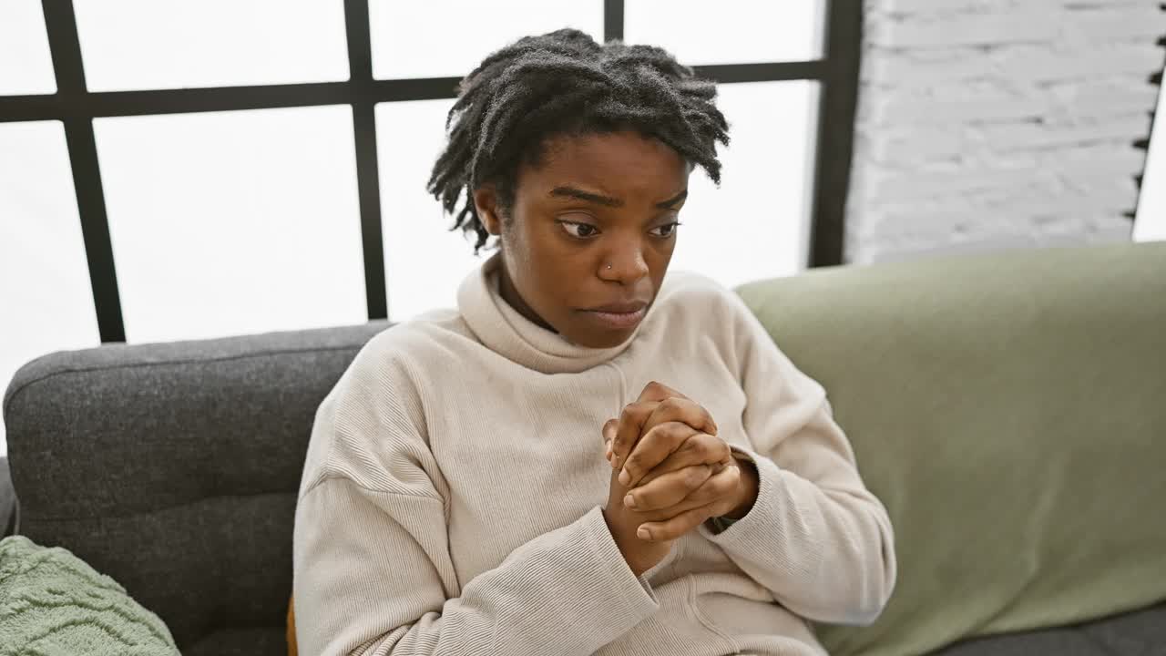 一个留着脏辫的沮丧的年轻黑人女子坐在沙发上，手指着上面——一个情绪化的非裔美国女性的不快乐的家庭肖像，带着悲伤和痛苦指明了方向视频下载