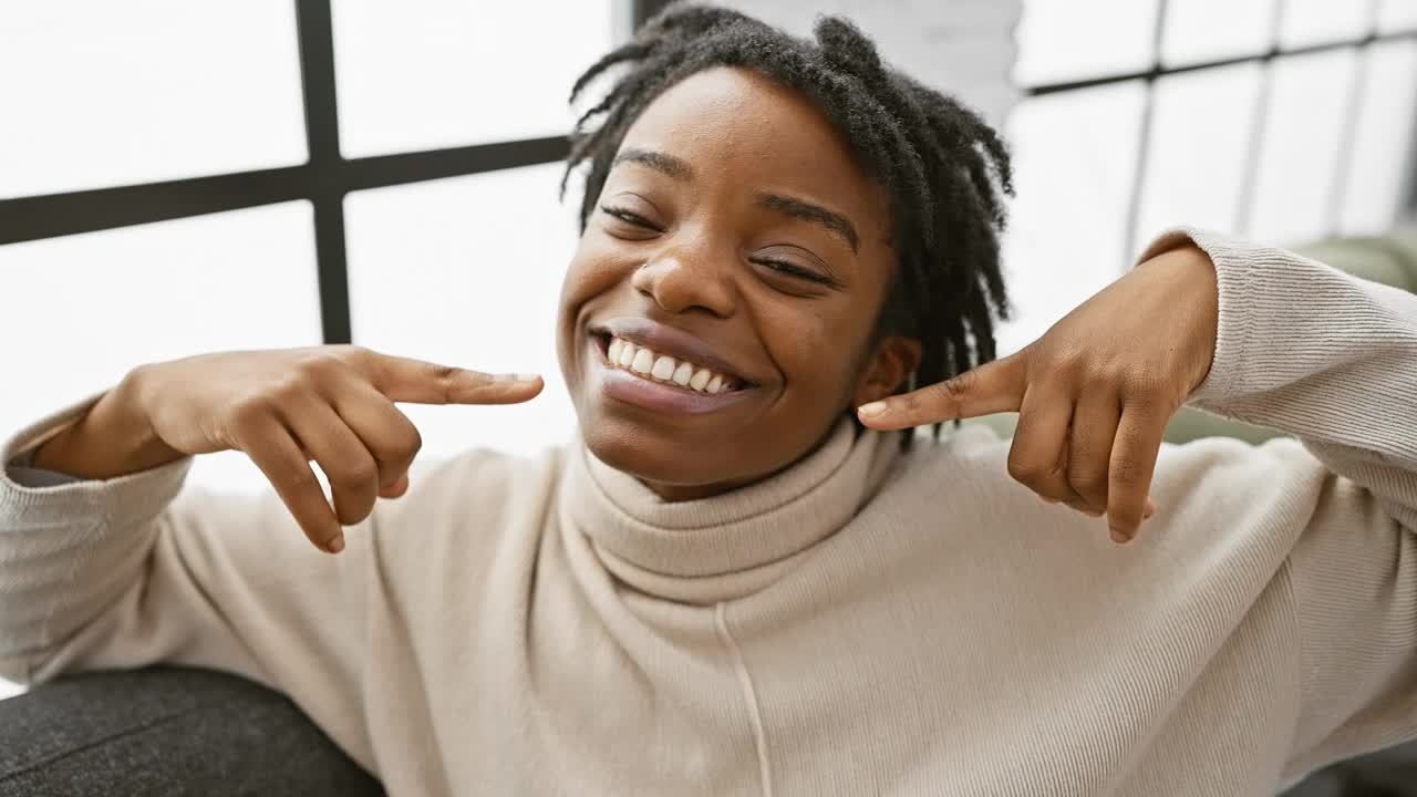 快乐的年轻黑人妇女，完美的牙齿闪闪发光，指着她健康的微笑舒适的客厅沙发，强调治疗成功的牙齿健康视频下载