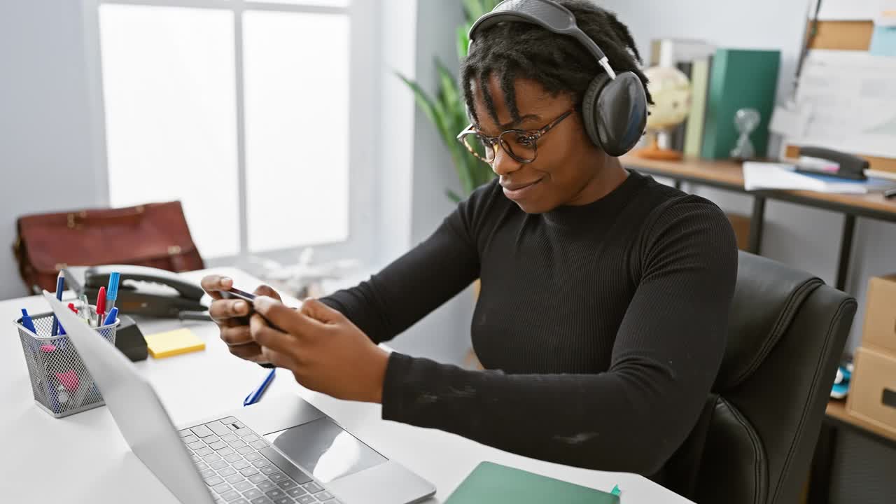 留着脏辫的非洲女性在办公室里用耳机和智能手机工作。视频下载