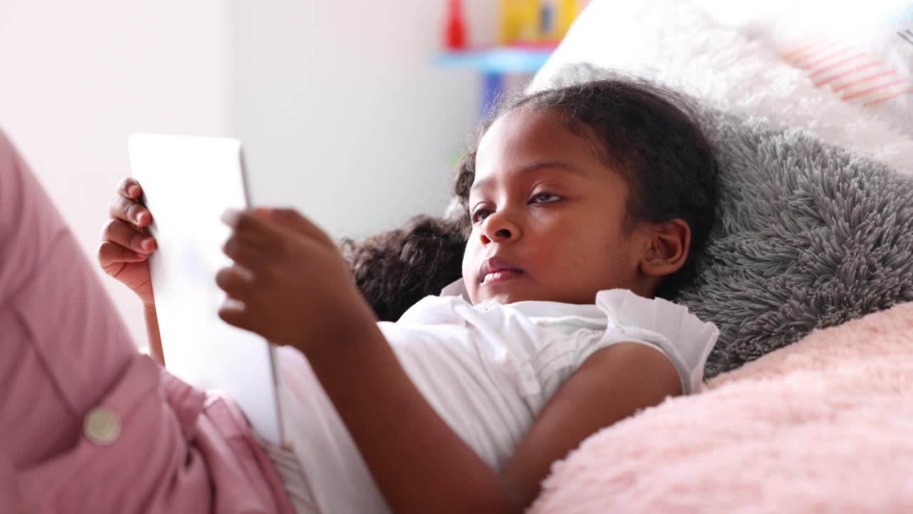 一个黑人小女孩躺在床上玩着数码平板电脑视频下载