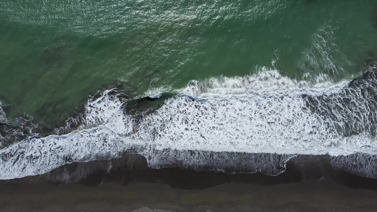 镜头向下看，海浪拍打着黑色的沙滩，深绿色的大海视频下载