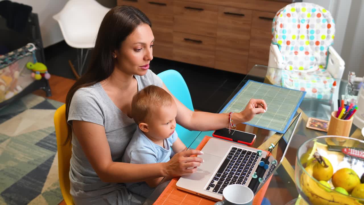 妈妈在看笔记本电脑上的视频，她的宝贝儿子坐在家里的腿上视频下载