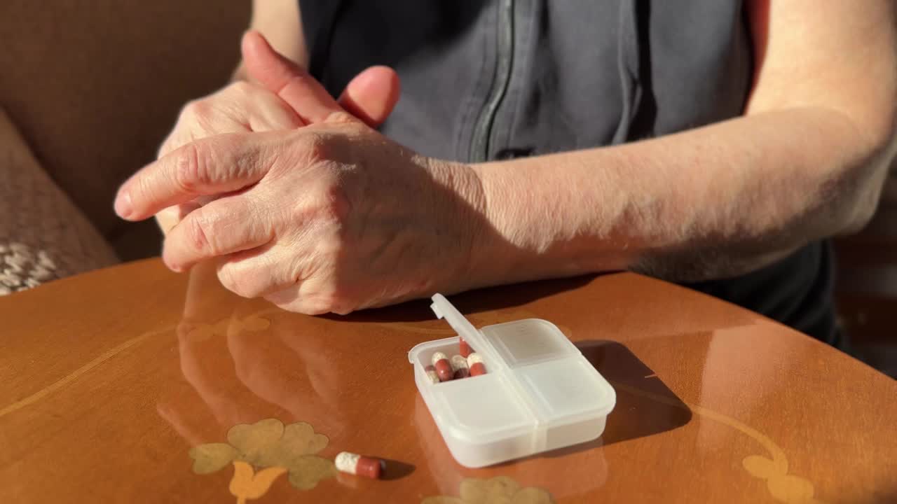 一位老妇人的手的特写，她的手紧握在一个装有药片的白色容器旁边视频下载