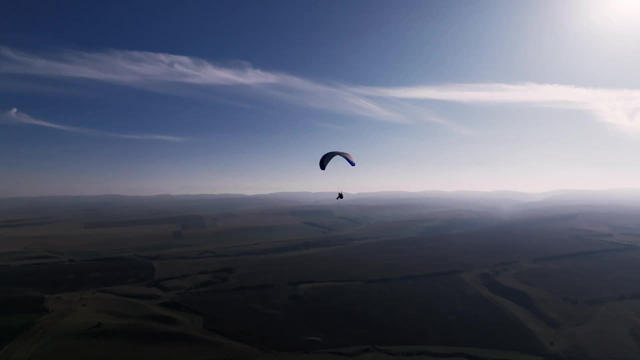 鸟瞰图，地平线上稳定的画面。一只雄性滑翔伞在田野和山丘的包围中飞行。深秋。蓝白色的滑翔伞翅膀在晴朗的天气视频下载