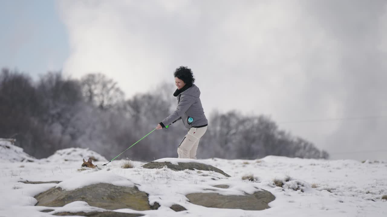 年轻女子带着法国斗牛犬漫步在山间冬日的雪景中，慢动作视频下载