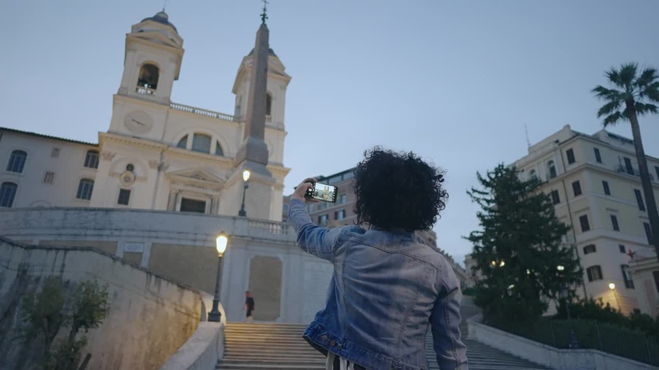 黄昏时分，一名西班牙卷发女子在罗马的西班牙阶梯上用智能手机拍照。电影慢动作镜头。视频下载