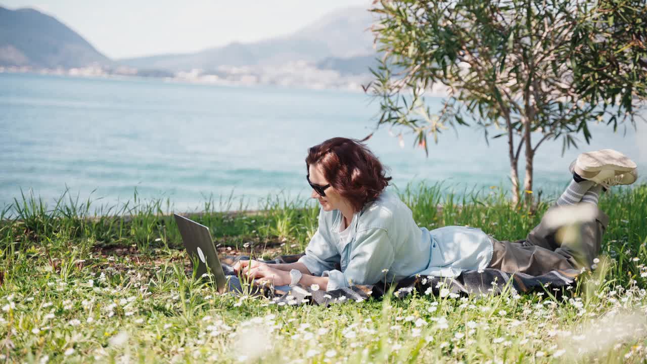 一位年轻女子躺在盛开的草坪上，正在用笔记本电脑工作视频下载