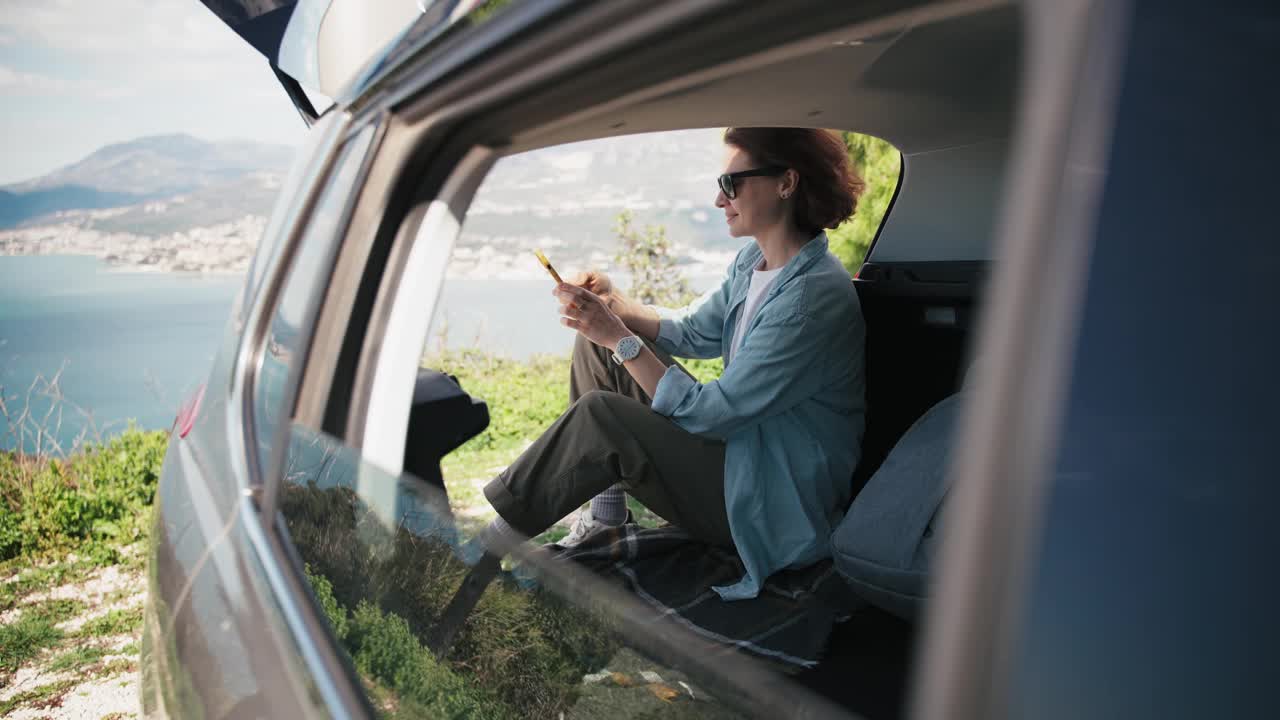一名年轻女子坐在汽车后备箱里玩智能手机视频下载