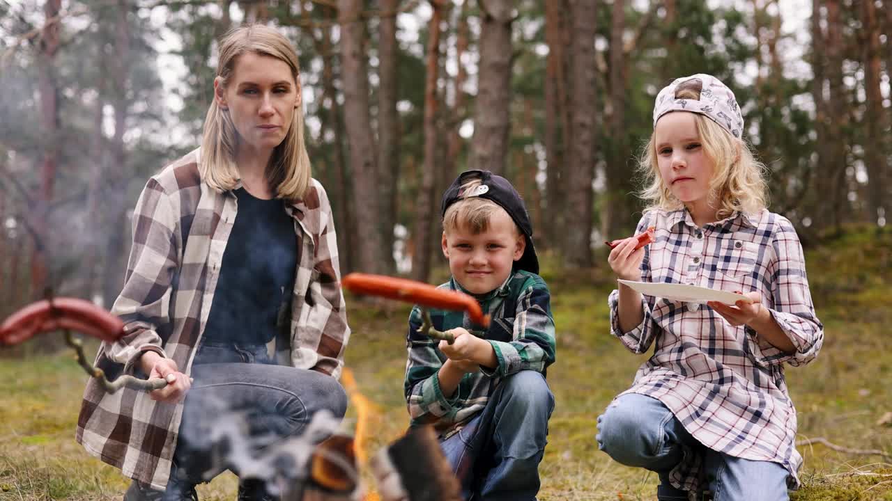 在森林里露营时，母亲带着孩子在篝火旁煎香肠吃。家庭时光，自然探险视频下载