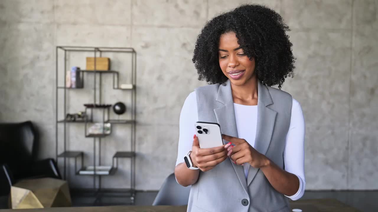 一位穿着时髦的灰色运动夹克的开朗的非裔美国妇女带着灿烂的微笑看着她的智能手机视频下载