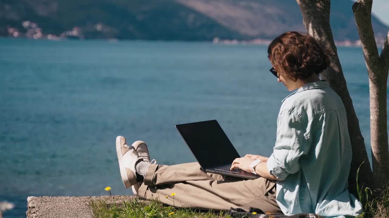 一位年轻的白人妇女坐在海边用笔记本电脑工作视频下载