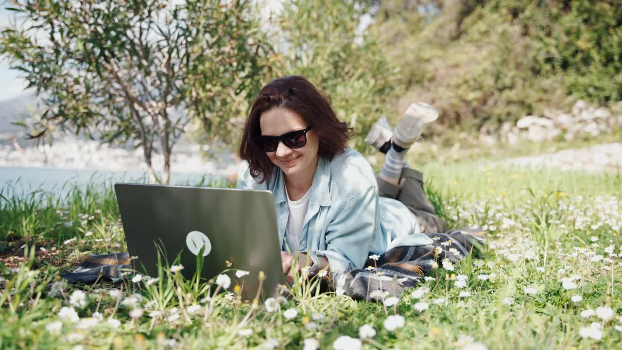 一位年轻女子躺在盛开的草坪上，正在用笔记本电脑工作视频下载