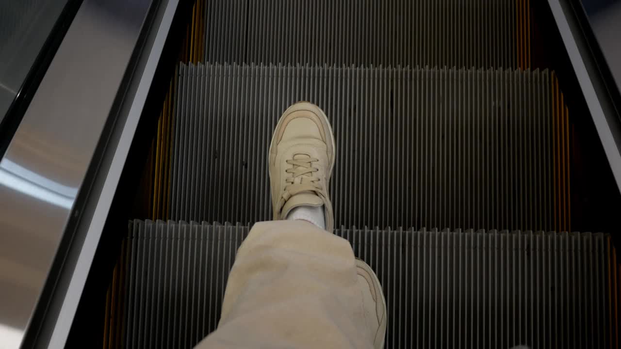 上面的特写显示了一个人的腿，一双穿着运动鞋的旅客在机场候机楼乘坐向下移动的自动扶梯。商场里自动扶梯扶手的俯视图。视频下载
