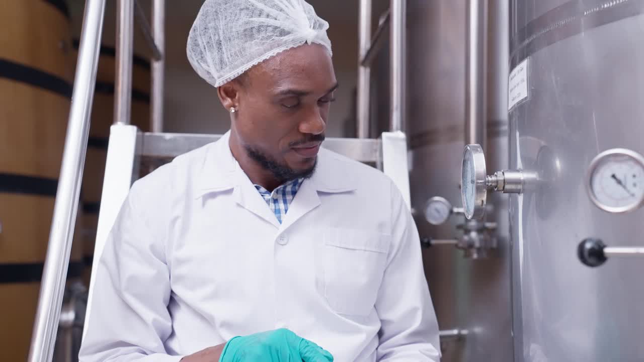 一位身穿实验室大褂、头戴发套的非洲成年男性主管，正在检查酒桶，检查发酵室里酒桶的压力，并在一台数字平板电脑上工作，以监控酿酒厂的质量。视频下载