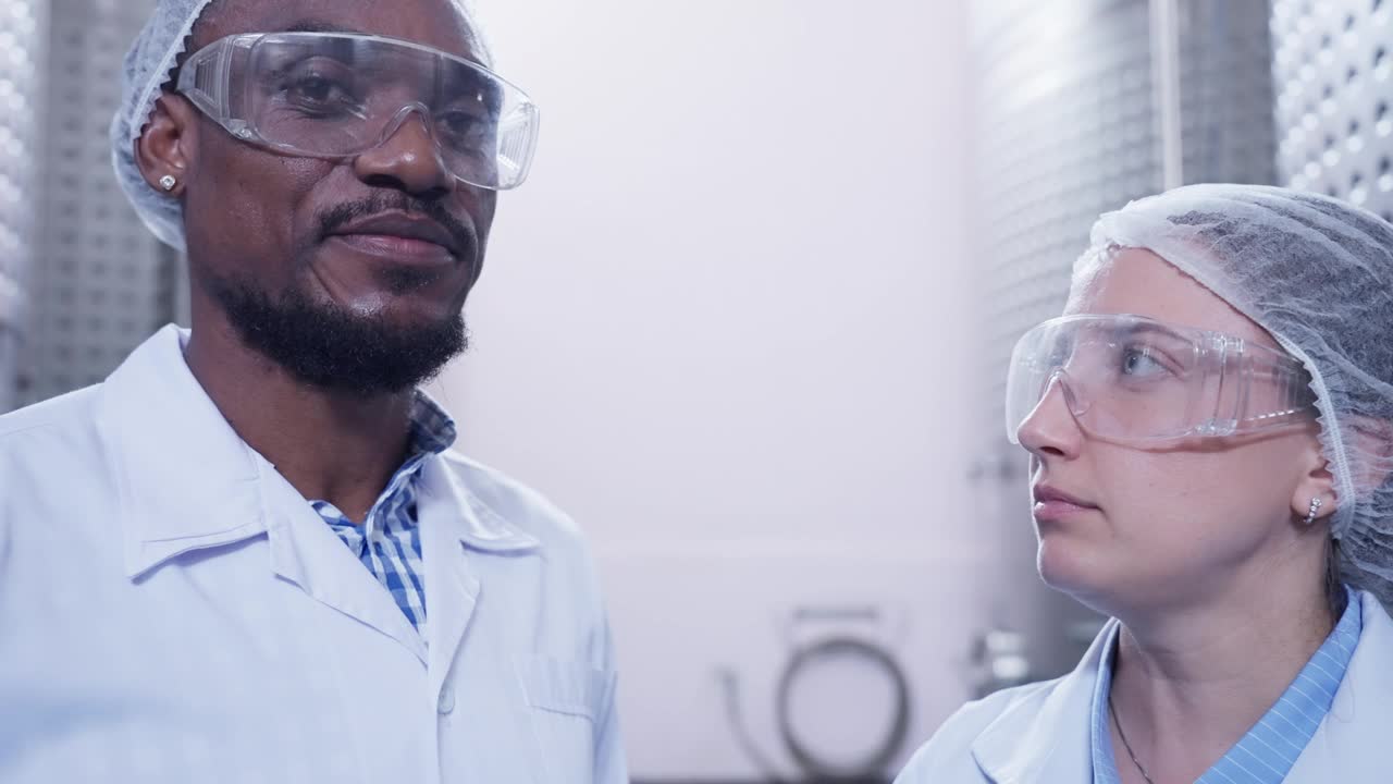 两位中年专家研究人员，非洲黑人男性和白人女性，都配备了实验室设备，微笑着站在葡萄酒发酵室里，周围都是不锈钢发酵罐。视频下载