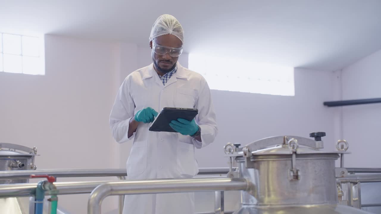 中年黑人男性酿酒师穿着洁净的实验室大褂，戴着发网，在容器发酵过程中仔细检查酒精饮料的质量，同时在数字平板上记录细节。视频下载