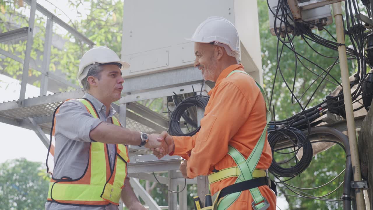 两名穿着防护服的男工程师同事在电信塔的控制面板前握手的特写镜头，反映了他们对完成工作的满意。视频下载