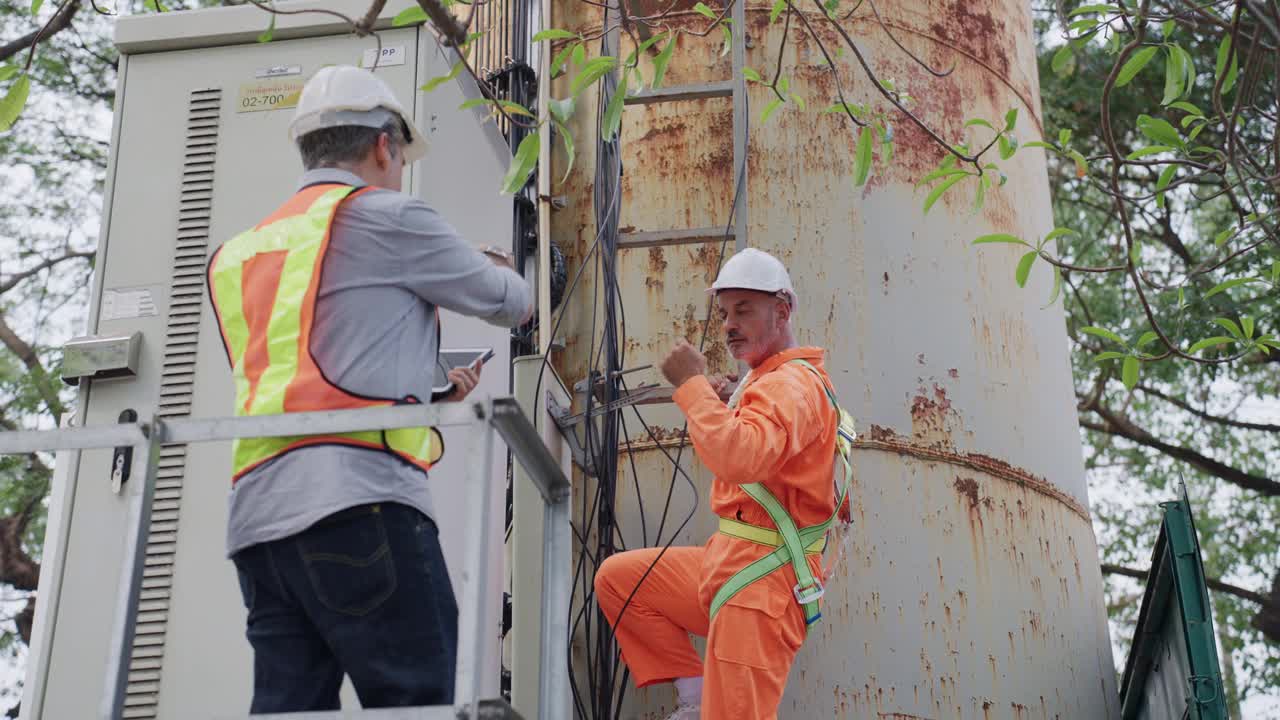 成熟的成年工程师穿着橙色防护服爬上变电站的梯子，他的同事用数码平板电脑与他沟通并检查站的性能。视频下载