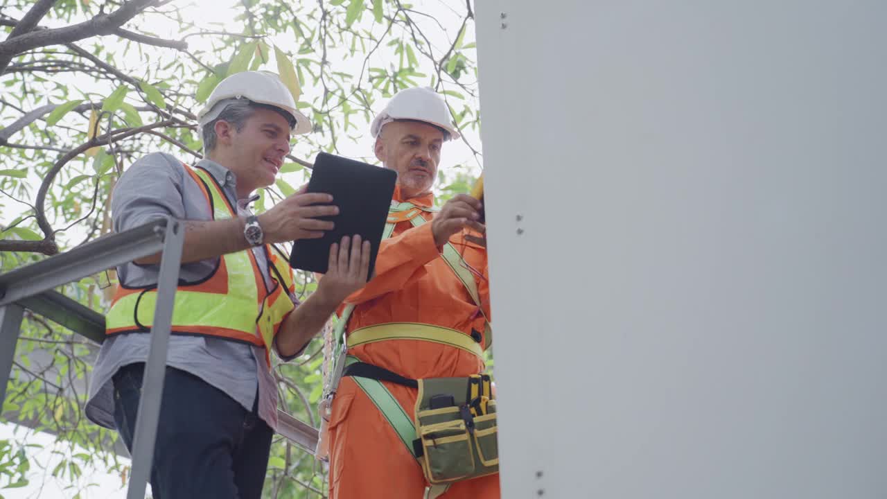 两名穿着防护服的成年男性技术人员站在电信塔前的特写镜头，一名技术人员使用电压表测量线路中的电流，而他的同事则在一台数字平板电脑上检查问题。视频下载