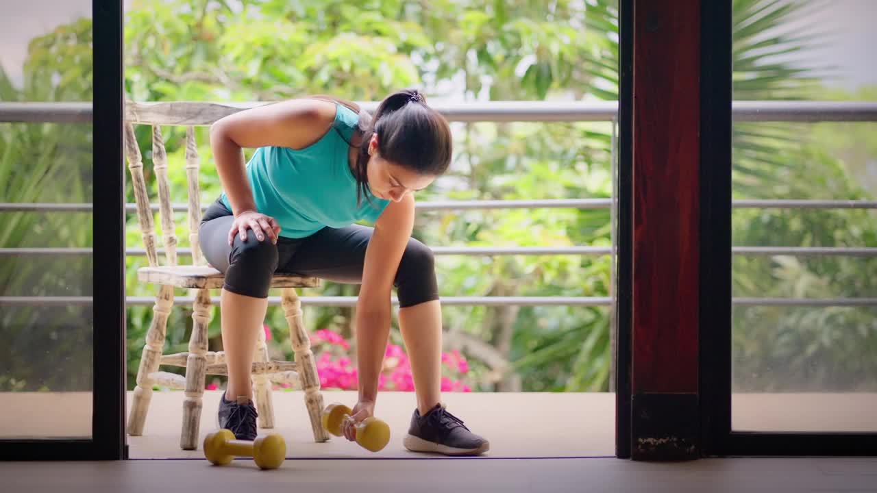 西班牙裔年轻女子在家里用哑铃健身坐在阳台的椅子上视频下载