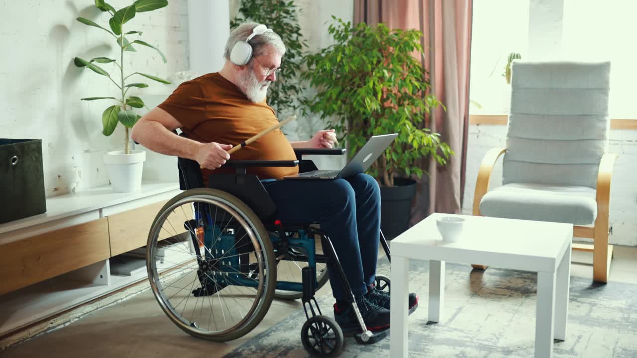 一位老人坐在轮椅上，戴着耳机，看着笔记本电脑，通过在线课程学习如何打鼓。室内设计视频下载