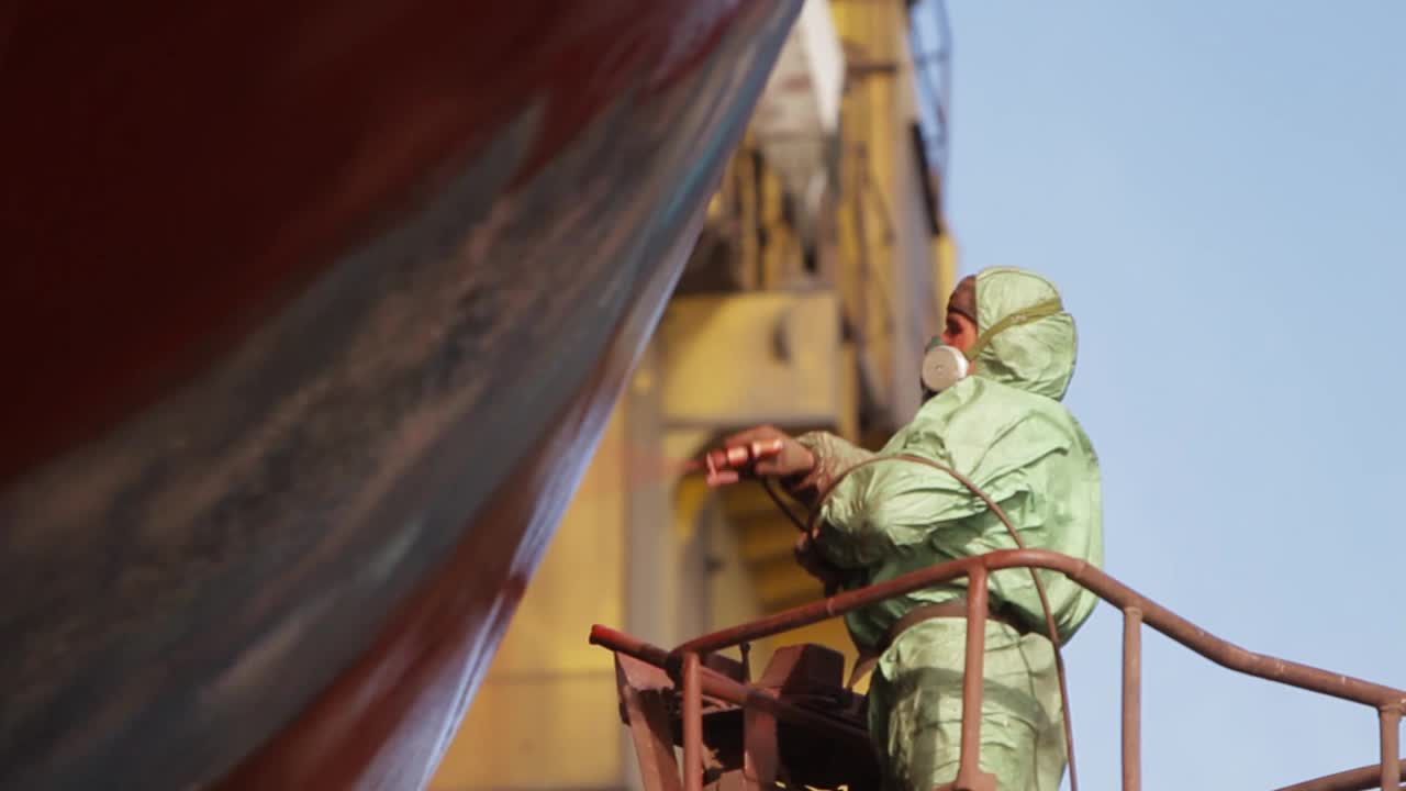 干船坞工人在船体上喷漆。专业的防护装备在船舶表面涂上红色涂层。海事维修、船舶工业现场。船厂劳动。视频下载
