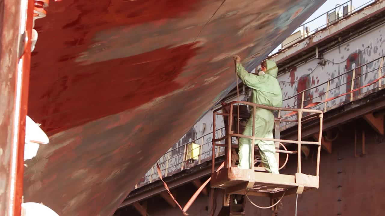 工人在船坞油漆船体，大型船只正在维修中。为安全而穿戴的防护装备，喷枪上涂有红色涂层。海运业，船舶改造过程在行动中展现。视频下载