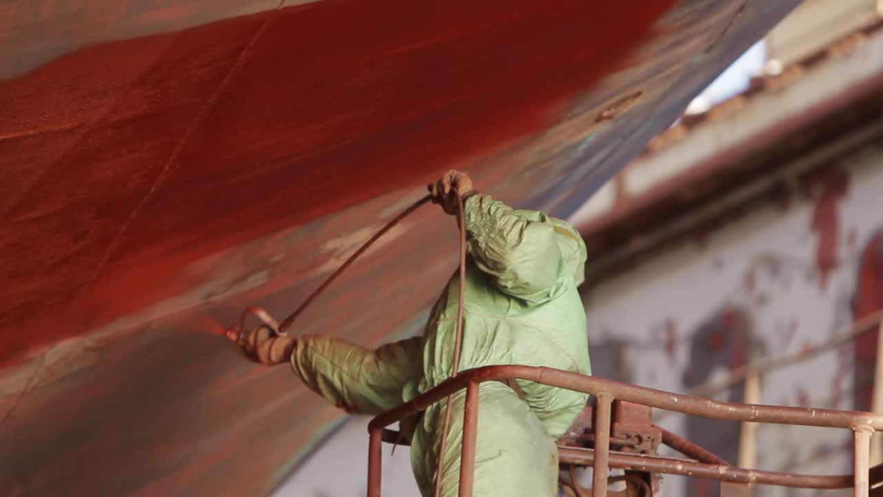 穿着防护服的工人在船厂给船体涂上红色油漆，翻新海运，船坞工人在行动，工业船舶维修，正在进行的海洋涂装过程。视频下载