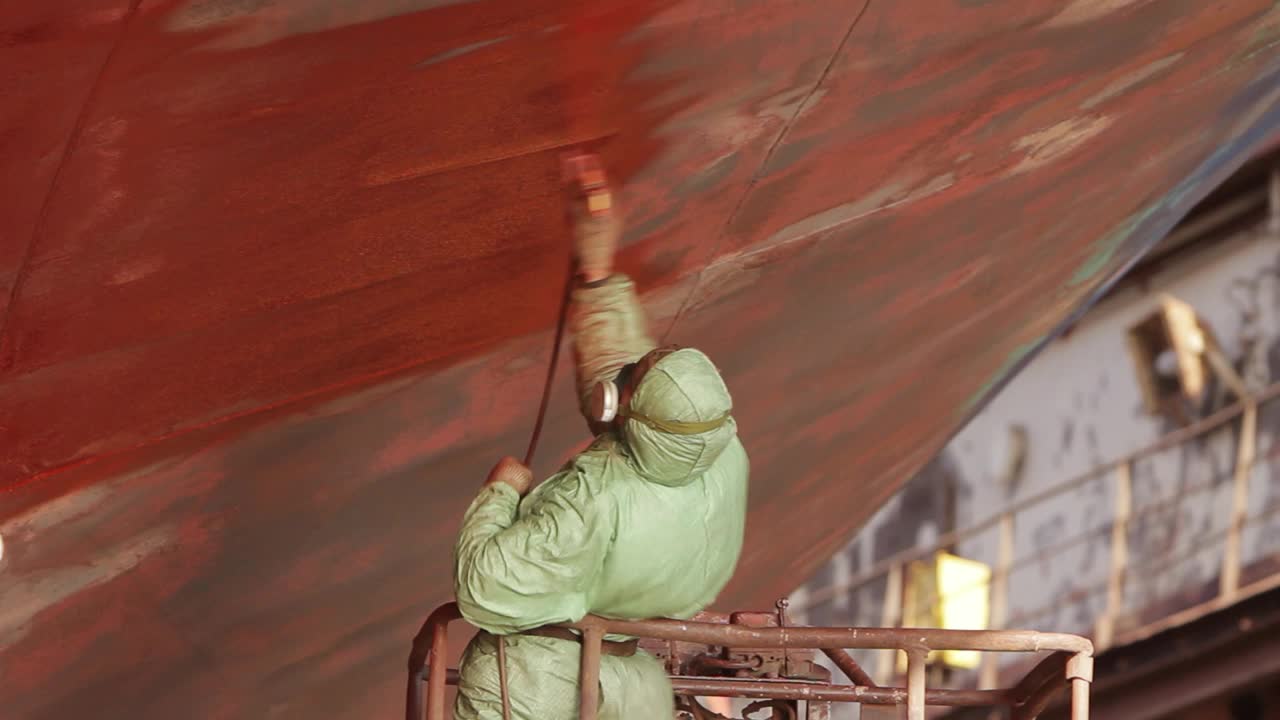 在干船坞，穿着防护装备的工人在船体上涂红漆。工业喷漆，防锈维修，船舶翻新工艺。船厂工人，使用安全设备。视频下载