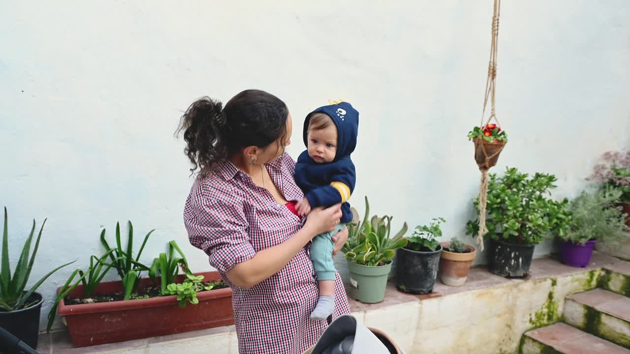 一个快乐的女人抱着她的小男孩，在她乡下房子的后院和他一起玩耍，房子里装饰着装饰性和烹饪用的草药和植物视频下载