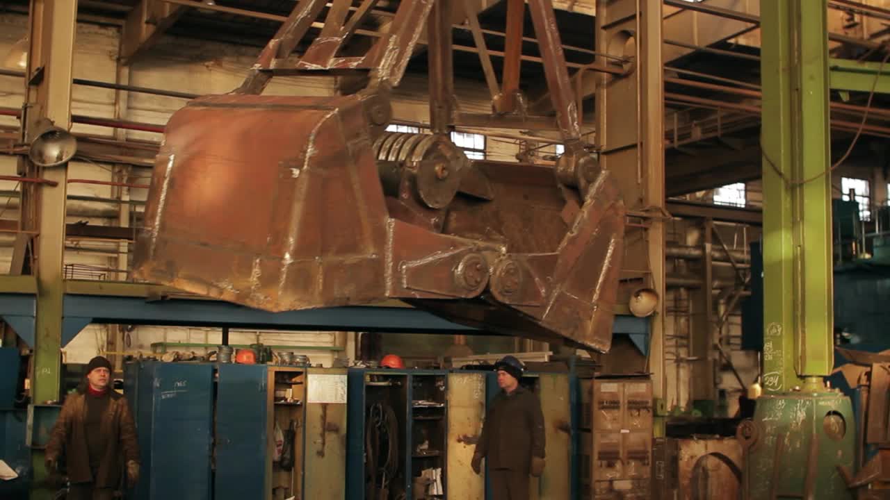 工人在机械厂操作工业起重机。吊车吊着钢包。戴着安全帽的工人在车间里引导重型设备。制造过程，团队合作。视频下载