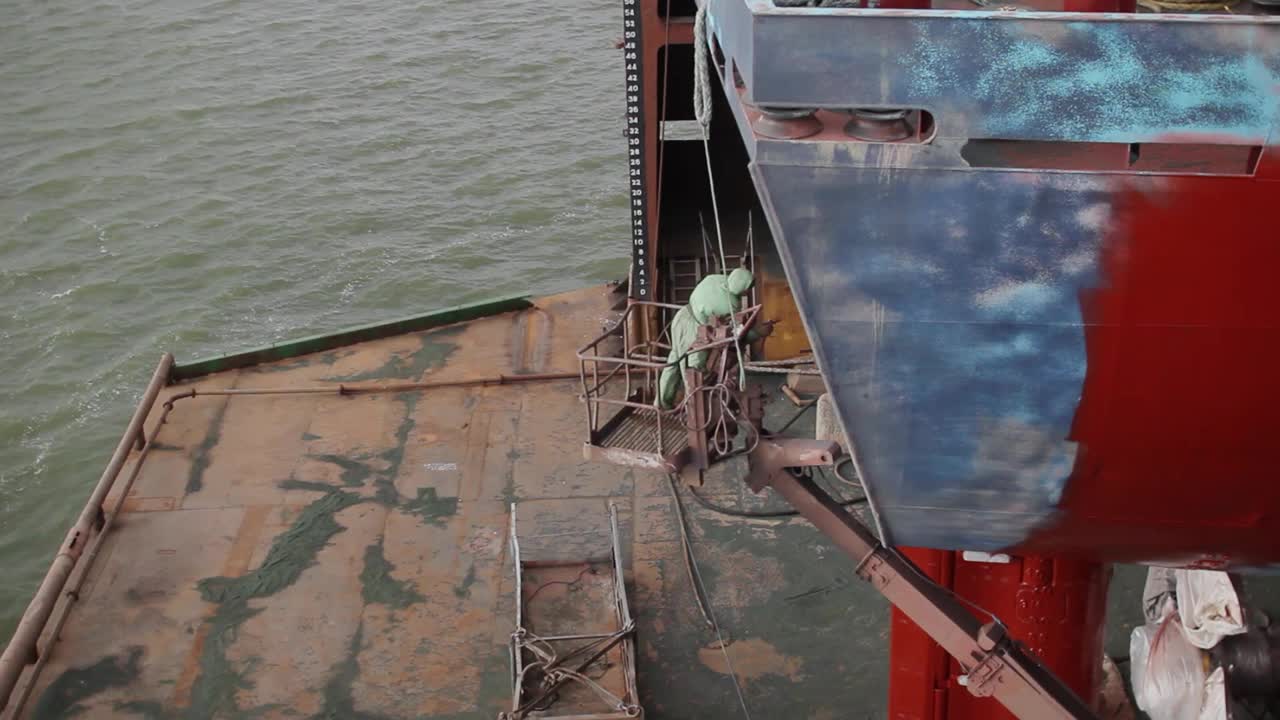 工人在干船坞给船壳上漆。工业船舶维修、除锈、货轮底漆应用。海事工作、船坞劳工、造船活动。俯视图，港口设施。视频下载