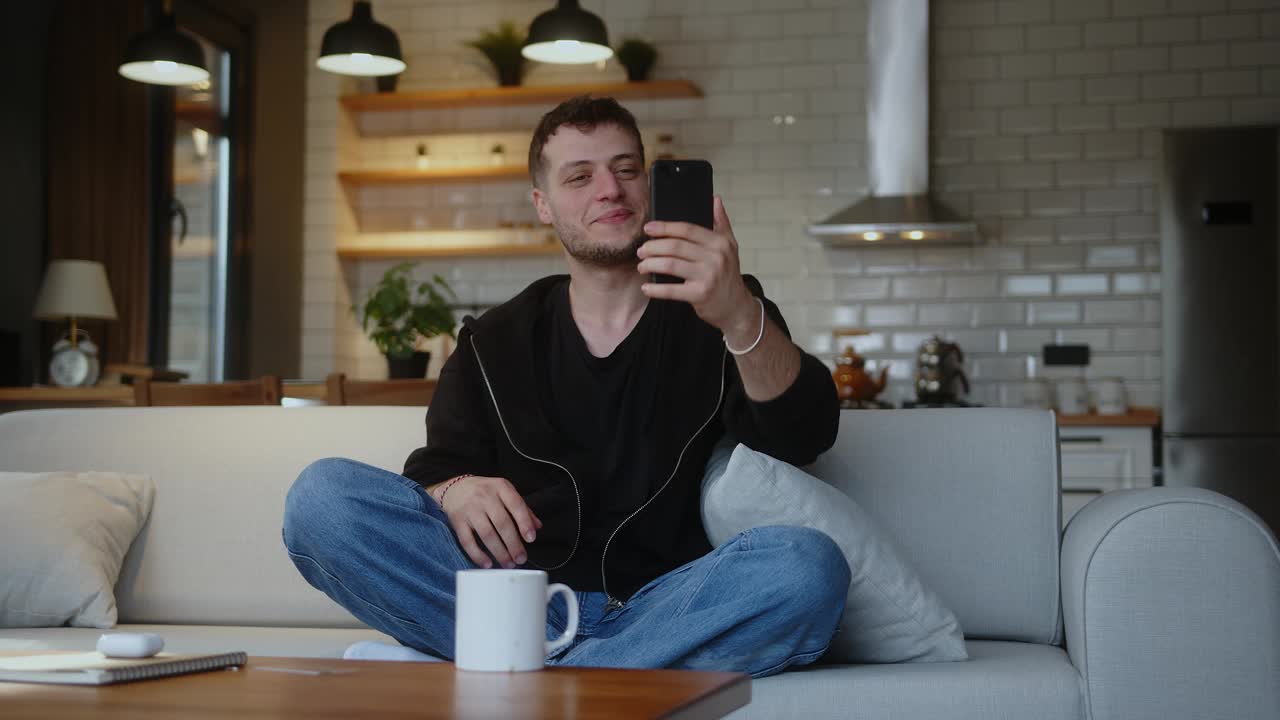 微笑的男子坐在沙发上拿着手机，一边用智能手机进行视频电话会议，一边挥手打招呼。男人看着屏幕，说话，微笑，在家享受在线聊天。视频下载