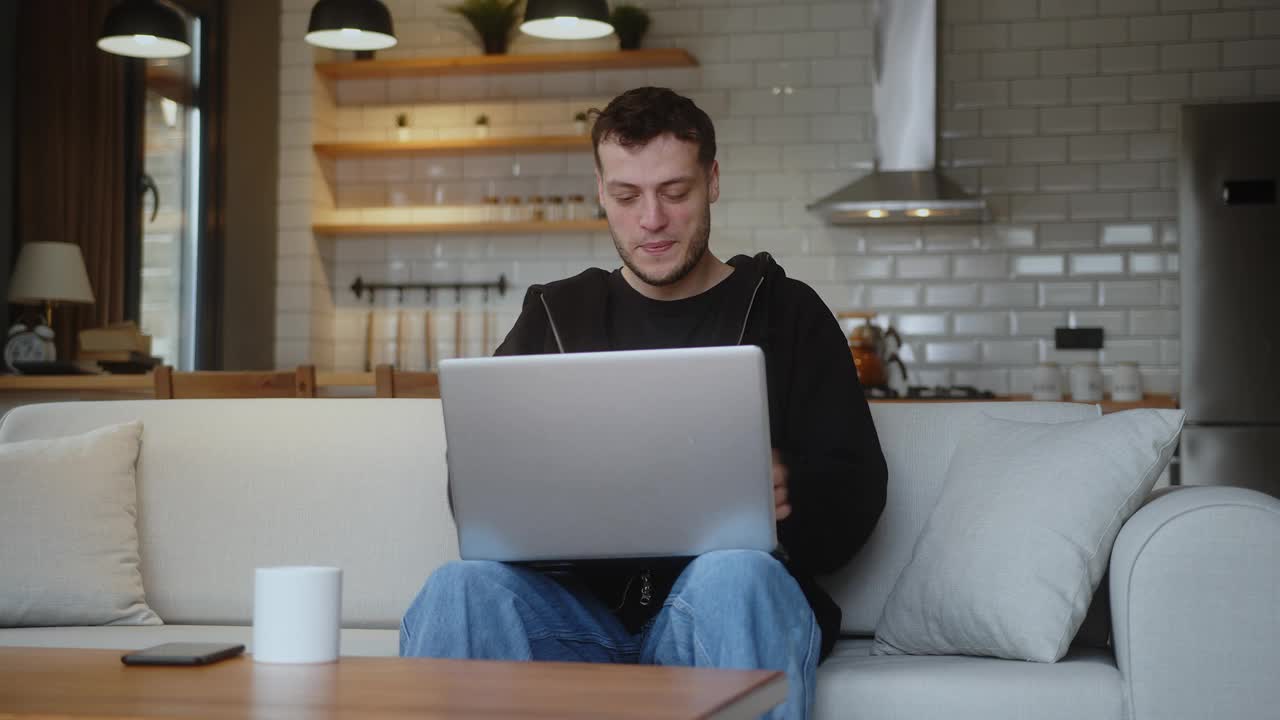 微笑的年轻人坐在客厅的沙发上用笔记本电脑工作。上网，用马克杯喝咖啡，在家里玩。视频下载
