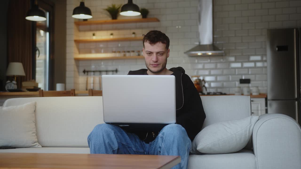 年轻人坐在沙发上用笔记本电脑看视频，上网，使用社交媒体，和朋友聊天。休闲活动，空闲时间在家客厅上网视频下载