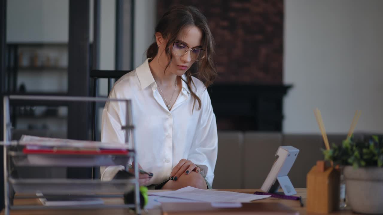 一位戴着眼镜、穿着白衬衫的女商人用笔填写了一份打印好的文件表格，并拿着桌上的一张撕下来的日历核对日期。一位女士坐在办公室的桌子旁视频下载